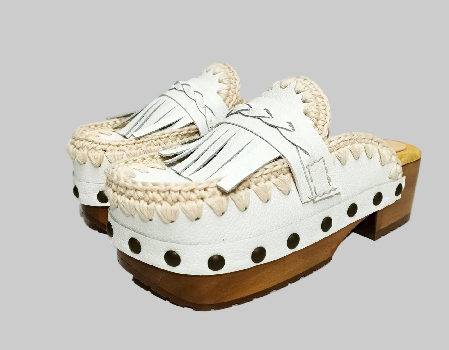 Handmade Niza Moccasin Clogs in white leather with beige crochet trim, leather fringes, and bronze studs on a wooden platform with a beige tractor sole.