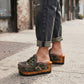 Detail shot of a model wearing Sol Caleyo's military camouflage platform clogs, showcasing the green and brown leather patterns, intricate crochet trim, and prominent antique gold hardware on an urban cobblestone background.