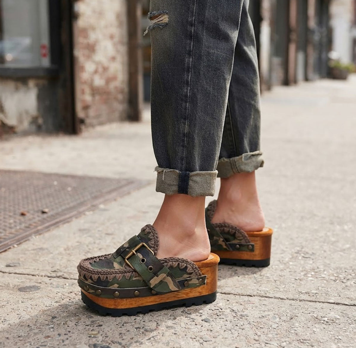 Detail shot of a model wearing Sol Caleyo's military camouflage platform clogs, showcasing the green and brown leather patterns, intricate crochet trim, and prominent antique gold hardware on an urban cobblestone background.