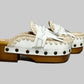 Handmade Niza Moccasin Clogs in white leather with beige crochet trim, leather fringes, and bronze studs on a wooden platform with a beige tractor sole.