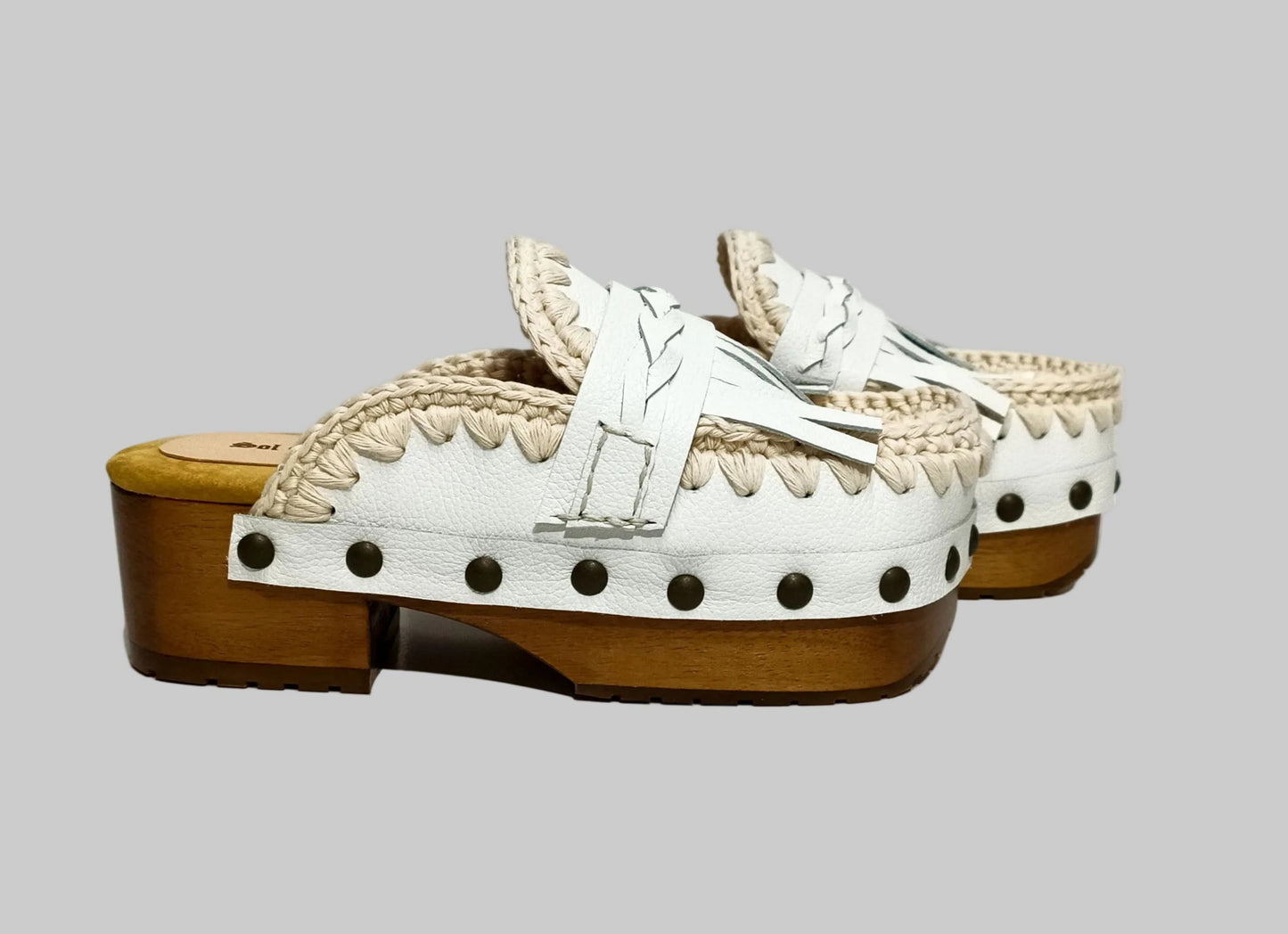 Handmade Niza Moccasin Clogs in white leather with beige crochet trim, leather fringes, and bronze studs on a wooden platform with a beige tractor sole.