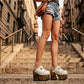 Close-up of a model wearing Sol Caleyo White Indian Clogs XL with 17cm heels and metal conchos, styled with denim shorts in an urban setting.