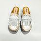 Handmade Niza Moccasin Clogs in white leather with beige crochet trim, leather fringes, and bronze studs on a wooden platform with a beige tractor sole.