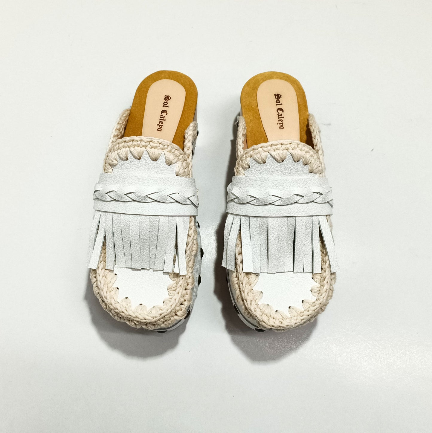 Handmade Niza Moccasin Clogs in white leather with beige crochet trim, leather fringes, and bronze studs on a wooden platform with a beige tractor sole.