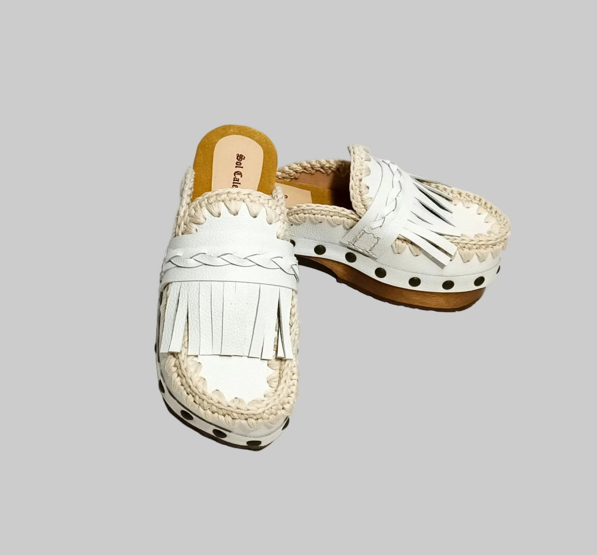 Handmade Niza Moccasin Clogs in white leather with beige crochet trim, leather fringes, and bronze studs on a wooden platform with a beige tractor sole.