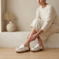 Woman wearing a flowing beige dress sits on a neutral stone ledge, displaying the Niza artisanal white leather platform clogs with braided leather straps and crochet trim.