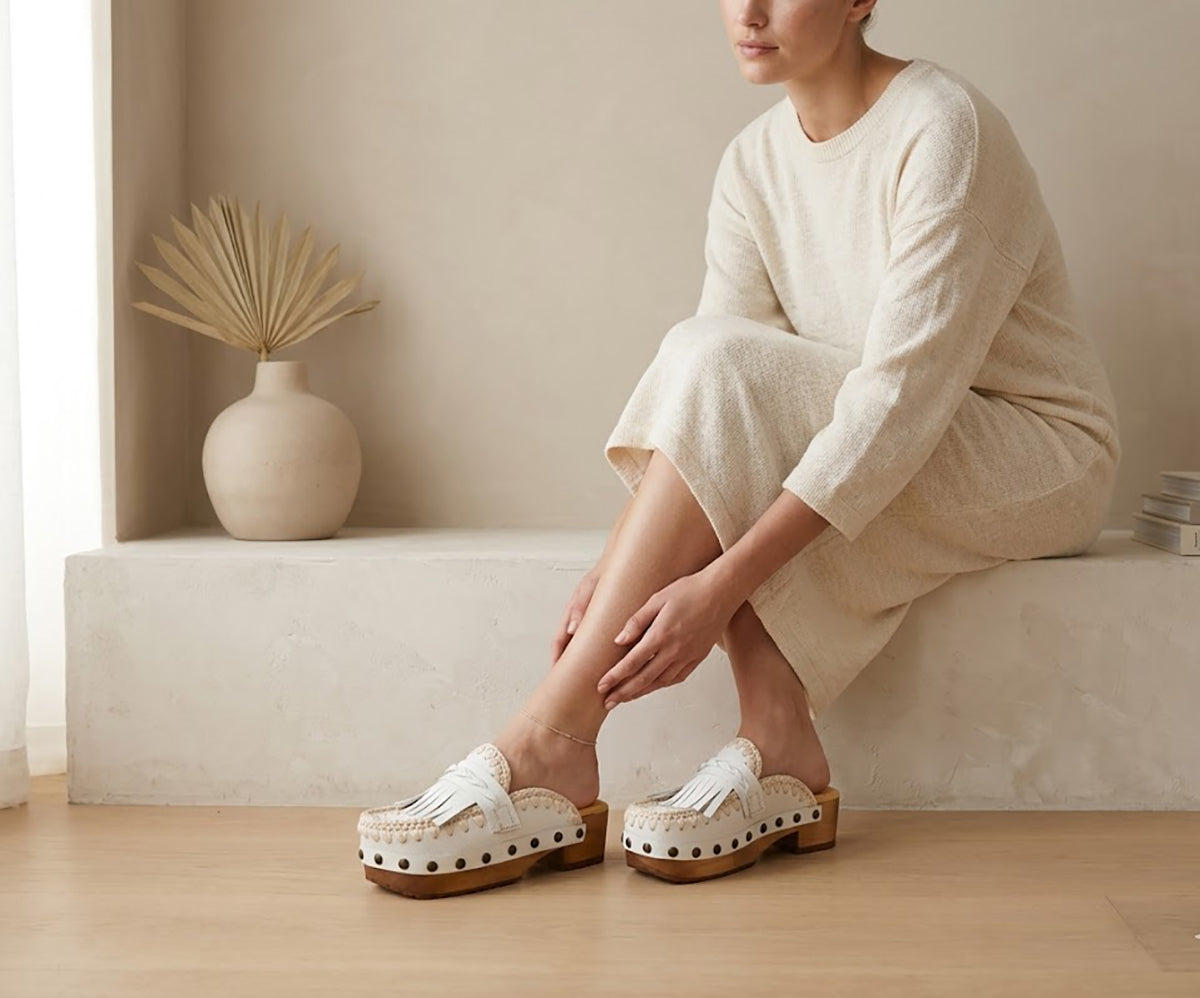 Woman wearing a flowing beige dress sits on a neutral stone ledge, displaying the Niza artisanal white leather platform clogs with braided leather straps and crochet trim.