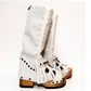 Side view of a pair of low-heeled Dakota boots in off-white suede against a white background. The boots feature a carved wooden platform sole and an open toe. They are decorated with fringe, a white wool braid down the side, and aged brass studs and rivets. A small charm hangs from the fringe on one ankle. The text ‘LOW HEEL’ appears in the lower right corner of the image.