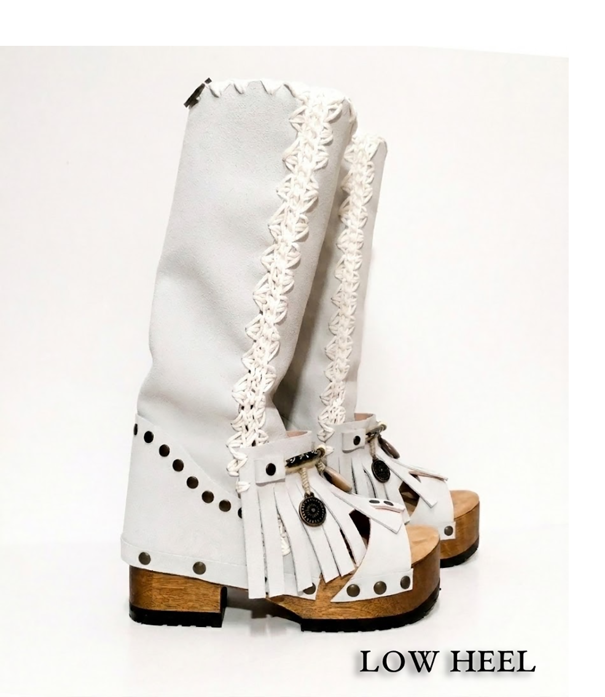 Side view of a pair of low-heeled Dakota boots in off-white suede against a white background. The boots feature a carved wooden platform sole and an open toe. They are decorated with fringe, a white wool braid down the side, and aged brass studs and rivets. A small charm hangs from the fringe on one ankle. The text ‘LOW HEEL’ appears in the lower right corner of the image.