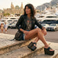 Woman sitting outdoors wearing Sol Caleyo Kauai XL platform flip flop sandals in black leather with silver studs, paired with a black leather jacket and denim shorts.