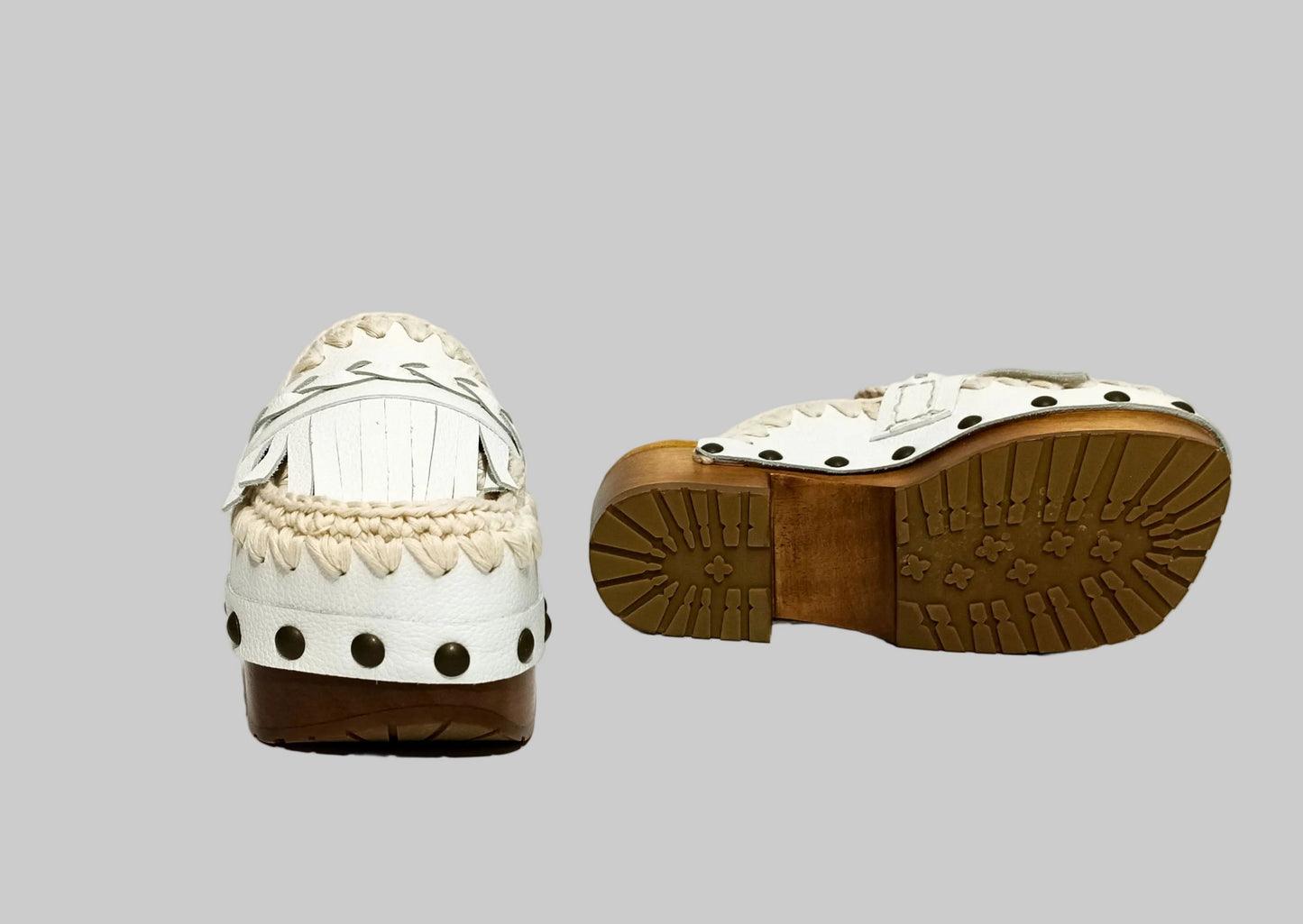Handmade Niza Moccasin Clogs in white leather with beige crochet trim, leather fringes, and bronze studs on a wooden platform with a beige tractor sole.