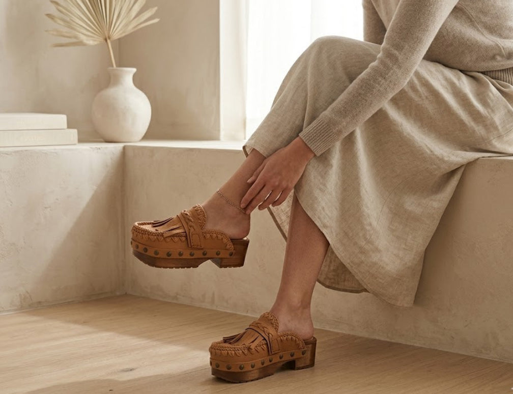 Close-up of the Niza tan leather platform clogs by Sol Caleyo, featuring hand-braided straps, fringes, and bronze studs on a carved wooden sole.