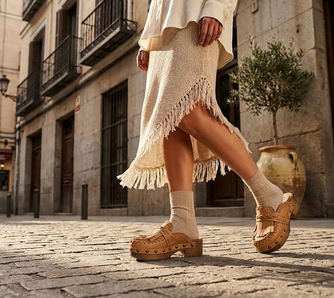 Sol Caleyo Niza Moccasin Clogs in light brown leather worn on a cobblestone street, featuring crochet details, fringes, and a wooden platform sole