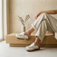 Close-up on legs wearing beige linen pants, showcasing the Niza handmade white leather moccasin clogs by Sol Caleyo with fringe details and a wooden platform sole.