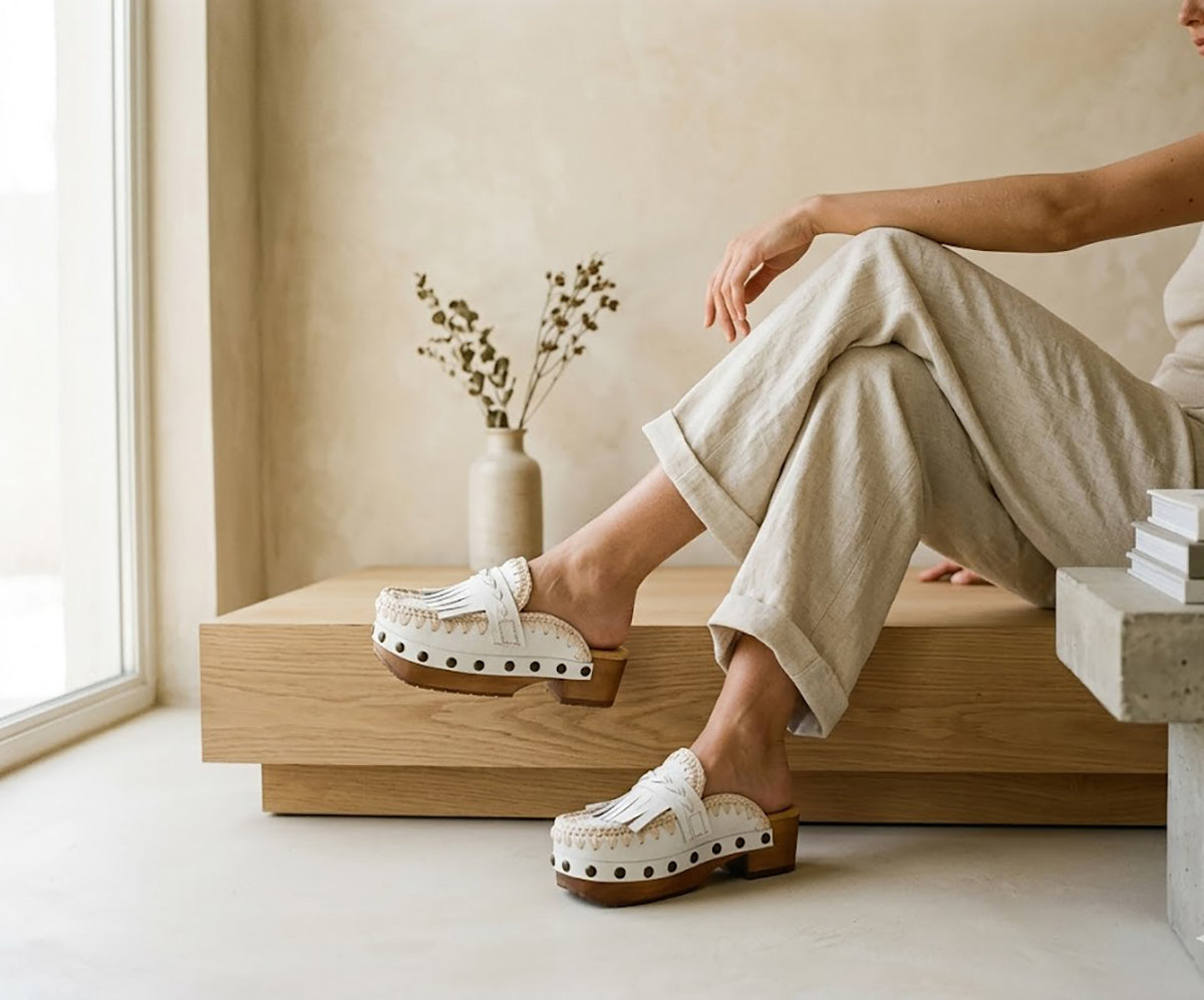 Close-up on legs wearing beige linen pants, showcasing the Niza handmade white leather moccasin clogs by Sol Caleyo with fringe details and a wooden platform sole.