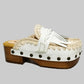 Handmade Niza Moccasin Clogs in white leather with beige crochet trim, leather fringes, and bronze studs on a wooden platform with a beige tractor sole.