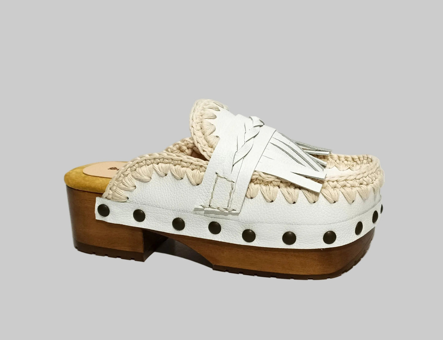Handmade Niza Moccasin Clogs in white leather with beige crochet trim, leather fringes, and bronze studs on a wooden platform with a beige tractor sole.