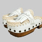 Handmade Niza Moccasin Clogs in white leather with beige crochet trim, leather fringes, and bronze studs on a wooden platform with a beige tractor sole.