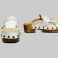 Handmade Niza Moccasin Clogs in white leather with beige crochet trim, leather fringes, and bronze studs on a wooden platform with a beige tractor sole.