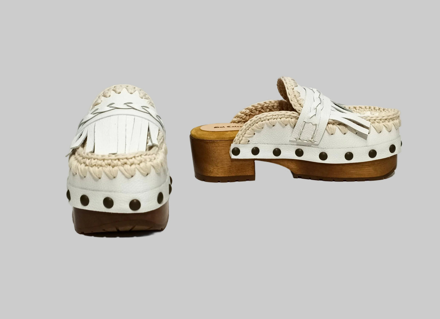 Handmade Niza Moccasin Clogs in white leather with beige crochet trim, leather fringes, and bronze studs on a wooden platform with a beige tractor sole.