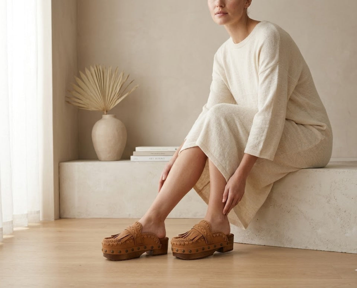 Close-up of the Niza tan leather platform clogs by Sol Caleyo, featuring hand-braided straps, fringes, and bronze studs on a carved wooden sole.