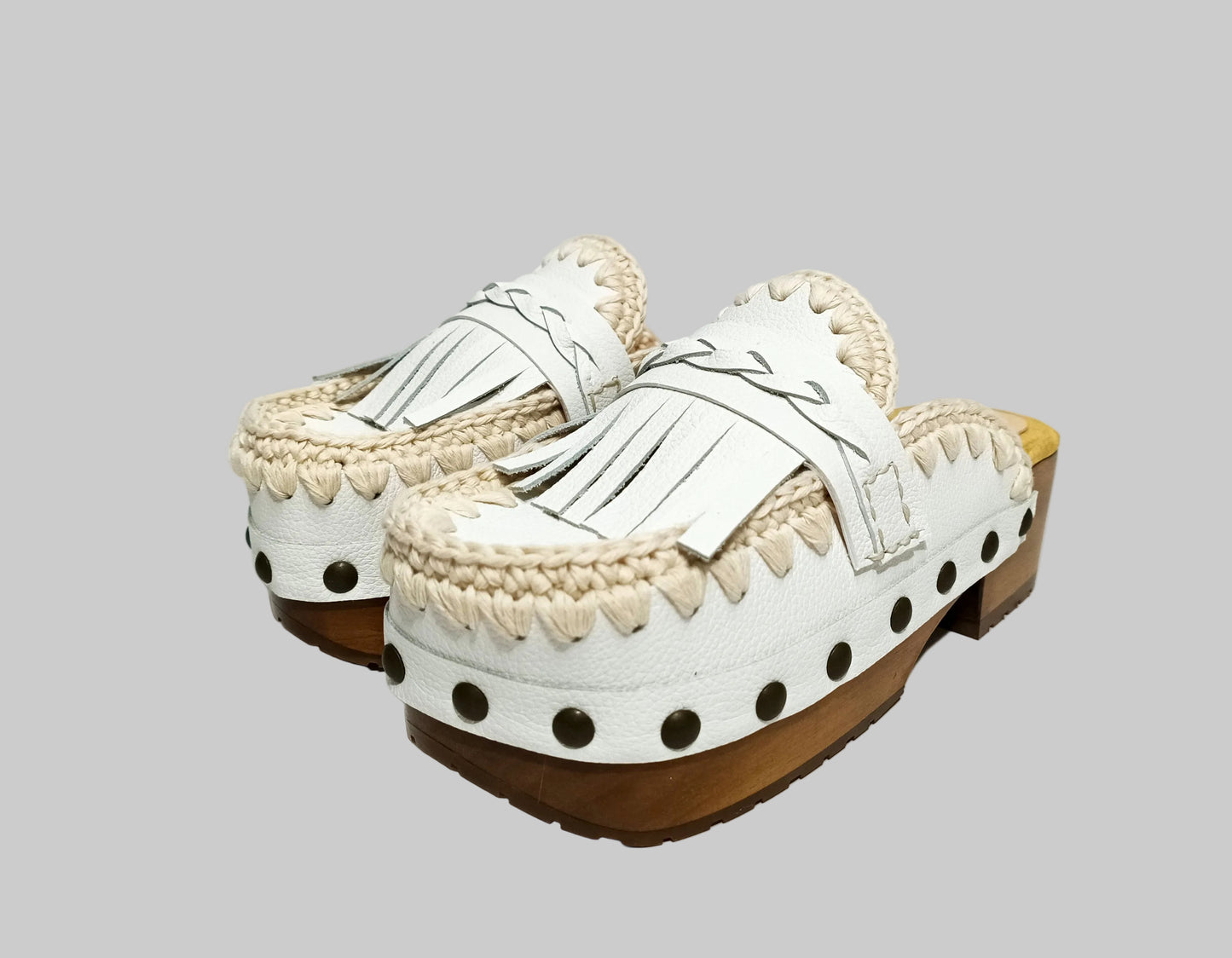 Handmade Niza Moccasin Clogs in white leather with beige crochet trim, leather fringes, and bronze studs on a wooden platform with a beige tractor sole.