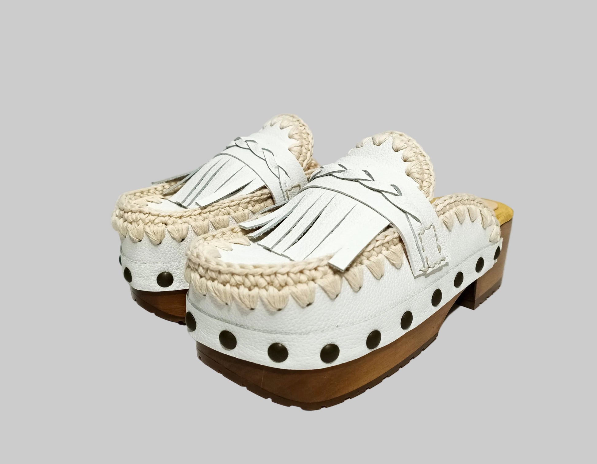 Handmade Niza Moccasin Clogs in white leather with beige crochet trim, leather fringes, and bronze studs on a wooden platform with a beige tractor sole.