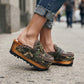 Detail shot of a model wearing Sol Caleyo's military camouflage platform clogs, showcasing the green and brown leather patterns, intricate crochet trim, and prominent antique gold hardware on an urban cobblestone background.