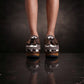 Person wearing Sol Caleyo's brown and white cow print leather clogs with tassels and woven trim on a platform, reflecting on a dark marble floor.