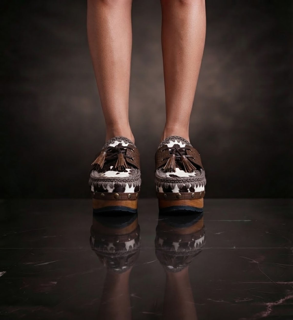 Person wearing Sol Caleyo's brown and white cow print leather clogs with tassels and woven trim on a platform, reflecting on a dark marble floor.