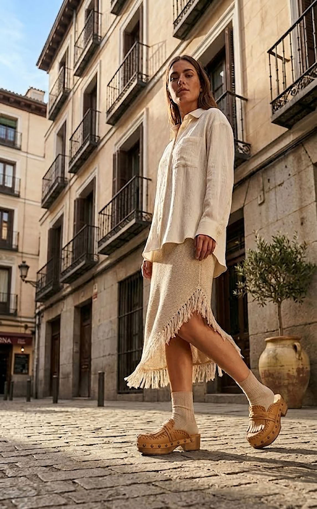 Sol Caleyo Niza Moccasin Clogs in light brown leather worn on a cobblestone street, featuring crochet details, fringes, and a wooden platform sole