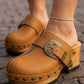 Close-up of a person wearing stylish light brown genuine leather clogs with an antique bronze buckle, wooden platform sole, and metallic studs, standing on a cobblestone street. Handmade designer footwear by Sol Caleyo.