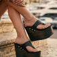 Close-up of a person's feet wearing Kauai XL black leather flip flop sandals with a high wengue wooden wedge platform and silver studs.
