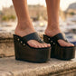 Close-up of a person's feet wearing Kauai XL black leather flip flop sandals with a high wengue wooden wedge platform and silver studs.