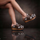 Person wearing Sol Caleyo's brown and white cow print leather clogs with tassels and woven trim on a platform, reflecting on a dark marble floor.