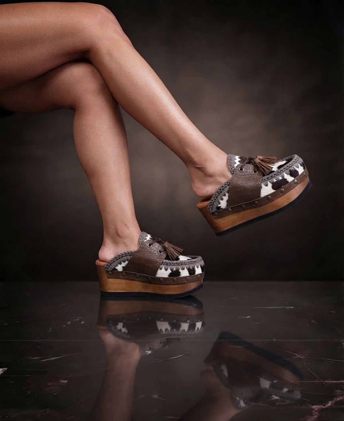 Person wearing Sol Caleyo's brown and white cow print leather clogs with tassels and woven trim on a platform, reflecting on a dark marble floor.