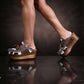 Person wearing Sol Caleyo's brown and white cow print leather clogs with tassels and woven trim on a platform, reflecting on a dark marble floor.