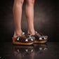 Person wearing Sol Caleyo's brown and white cow print leather clogs with tassels and woven trim on a platform, reflecting on a dark marble floor.