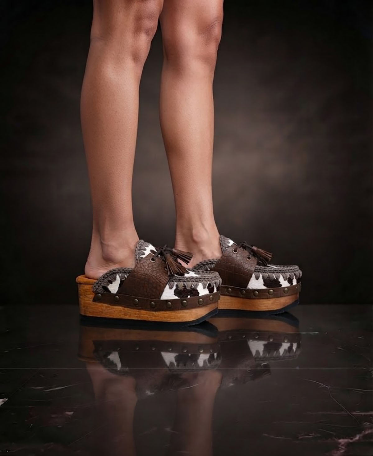 Person wearing Sol Caleyo's brown and white cow print leather clogs with tassels and woven trim on a platform, reflecting on a dark marble floor.
