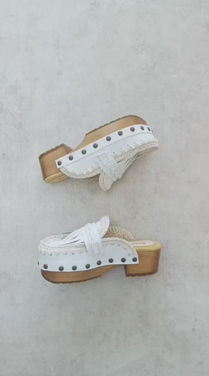 Handmade Niza Moccasin Clogs in white leather with beige crochet trim, leather fringes, and bronze studs on a wooden platform with a beige tractor sole.