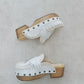 Handmade Niza Moccasin Clogs in white leather with beige crochet trim, leather fringes, and bronze studs on a wooden platform with a beige tractor sole.