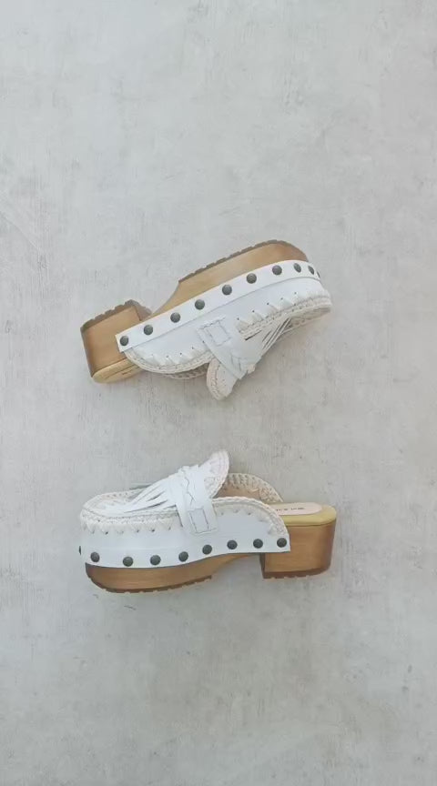 Handmade Niza Moccasin Clogs in white leather with beige crochet trim, leather fringes, and bronze studs on a wooden platform with a beige tractor sole.