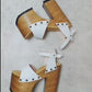 Side view of Crazy White Sandals by Sol Caleyo, featuring white leather straps with bronze studs on a high dark wood platform and tractor sole.
