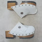 Top view of a pair of handmade white textured full-grain leather clogs with leather fringes and detailed Native American chief profile metal conchos on a wooden platform.