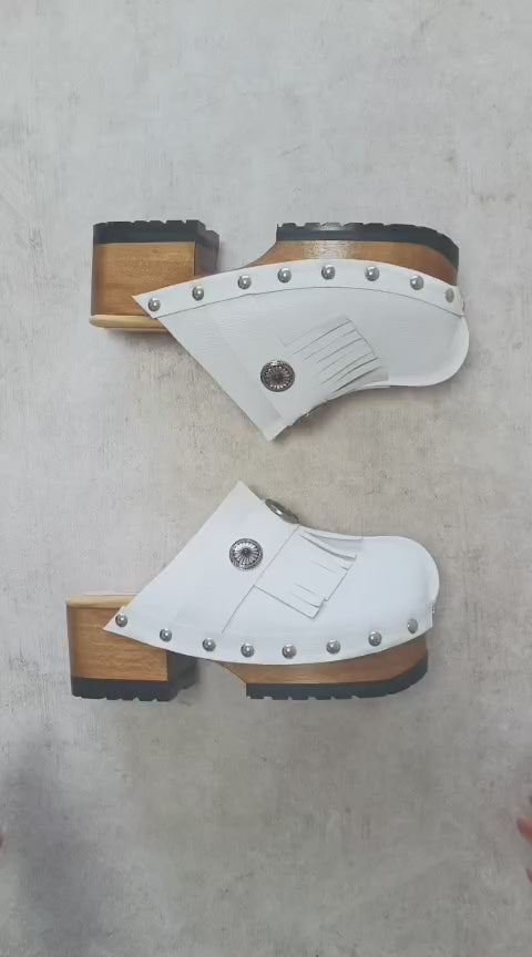 Top view of a pair of handmade white textured full-grain leather clogs with leather fringes and detailed Native American chief profile metal conchos on a wooden platform.