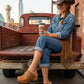 A woman wearing a cowboy hat, denim outfit, and Sol Caleyo handmade light brown leather Route 66 clogs with wooden platform soles, sitting in the back of a vintage red Chevrolet truck in an urban setting.