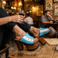 Close-up of a woman's legs wearing Sol Caleyo Argentina Sandals while relaxing at a rustic bar table in San Telmo. The wooden platform heels with the blue, white, and gold star Argentine flag design are clearly visible. She holds a glass of red wine.