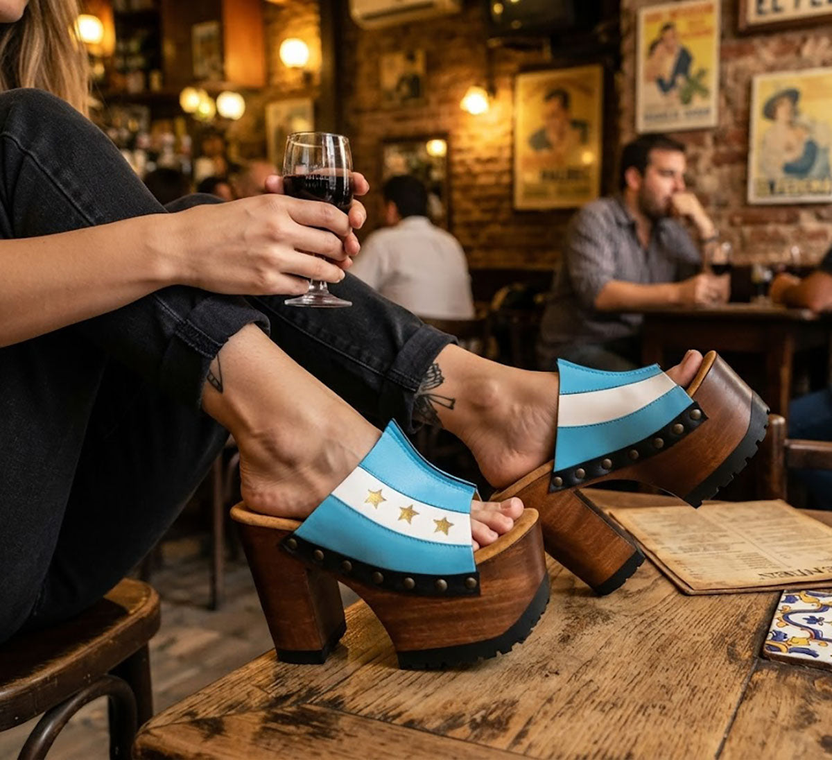 Close-up of a woman's legs wearing Sol Caleyo Argentina Sandals while relaxing at a rustic bar table in San Telmo. The wooden platform heels with the blue, white, and gold star Argentine flag design are clearly visible. She holds a glass of red wine.