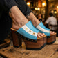 Close-up of a woman's legs wearing Sol Caleyo Argentina Sandals while relaxing at a rustic bar table in San Telmo. The wooden platform heels with the blue, white, and gold star Argentine flag design are clearly visible. She holds a glass of red wine.