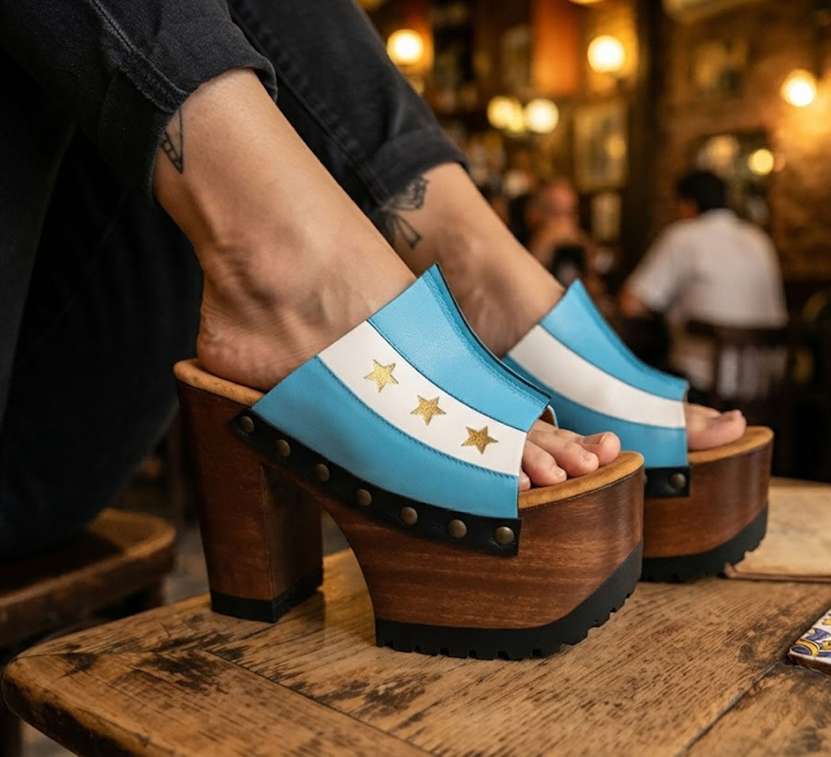 Close-up of a woman's legs wearing Sol Caleyo Argentina Sandals while relaxing at a rustic bar table in San Telmo. The wooden platform heels with the blue, white, and gold star Argentine flag design are clearly visible. She holds a glass of red wine.